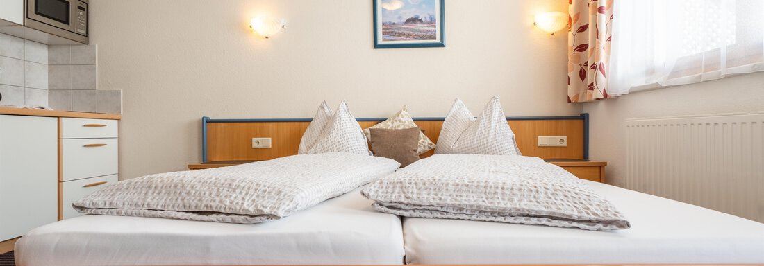 A cozy bedroom with a double bed and two pillows. In the background, there is a kitchen corner and a window with curtains. - Ferienwohnung Romana - Obergurgl 