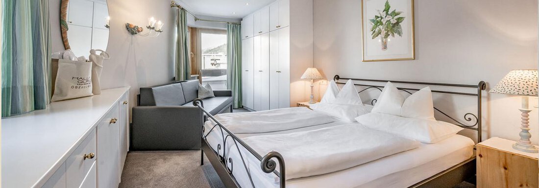 Ein helles Schlafzimmer mit einem Doppelbett und weißen Möbeln. Im Hintergrund befindet sich eine Sitzecke und große Fenster, die Tageslicht hereinlassen. - Skihotel Haus Gurgl - Obergurgl 
