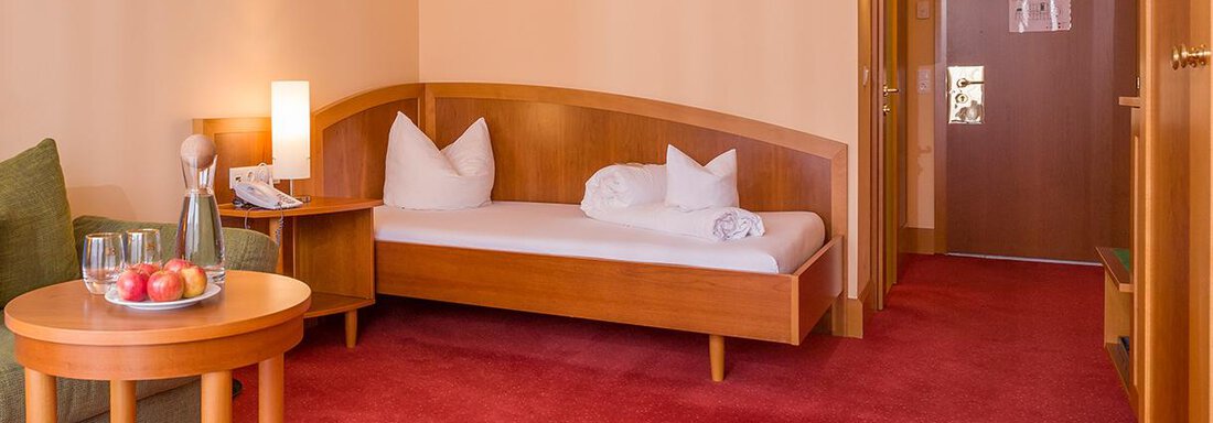A simple room with a red carpet and wooden furniture. There is a couch, a table with fresh fruit, and a single bed with pillows. - Vivea 4* Hotel Umhausen im Ötztal - Umhausen