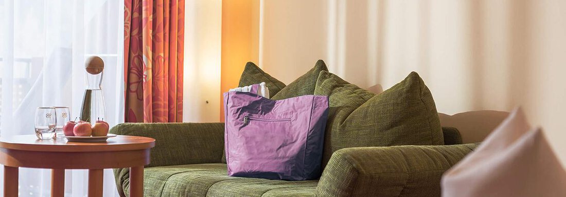 A cozy living room with a green sofa and colorful cushions. A small table with glasses and fruit stands next to it, and the light is bright and inviting. - Vivea 4* Hotel Umhausen im Ötztal - Umhausen