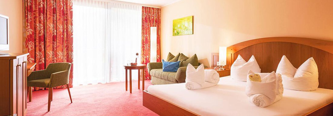 Vivea Hotel Umhausen double room | © Hannes Dabernig (CC BY) A cozy hotel room with a large bed and colorful pillows. The room has large windows, a table and chairs, as well as a television. - Vivea 4* Hotel Umhausen im Ötztal - Umhausen