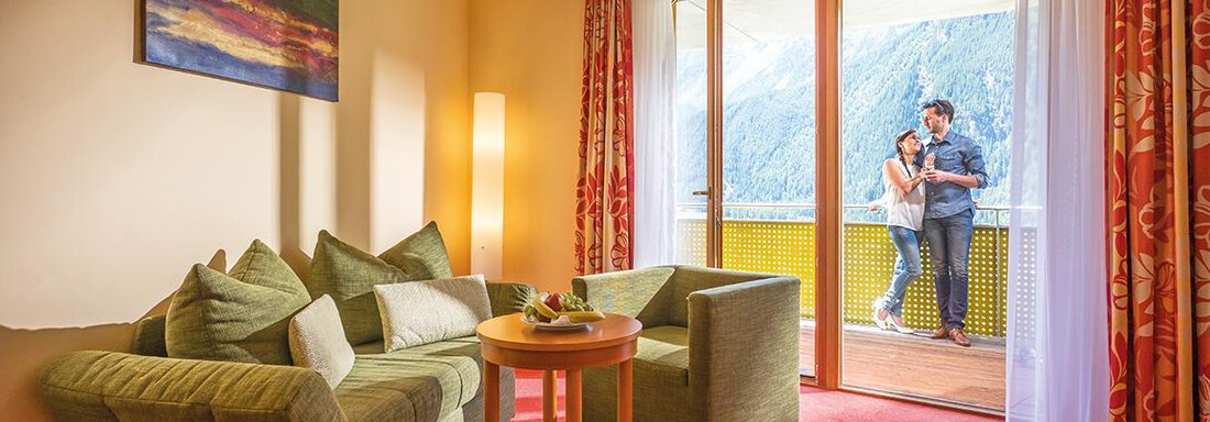 Vivea Hotel Umhausen suite | © Hannes Dabernig (CC BY) A cozy room with a seating area and large windows that offer a view of the mountains. A couple stands on the balcony and enjoys the view. - Vivea 4* Hotel Umhausen im Ötztal - Umhausen