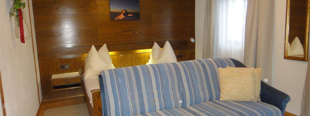 A cozy room with a blue sofa and a large bed with two pillows. The walls are made of wood, and a picture hangs above the bed. - Haus Elisabeth - Vent