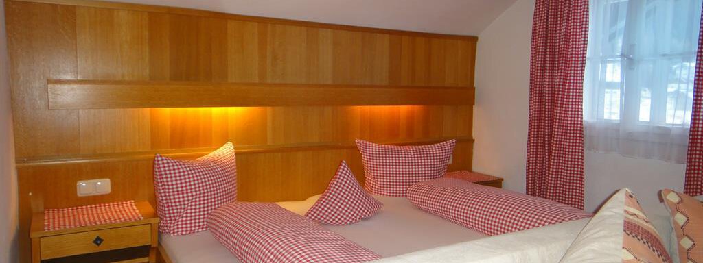 A cozy bedroom with wooden shelves and red-and-white checkered pillows. There is a large bed and windows with white curtains. - Haus Elisabeth - Vent