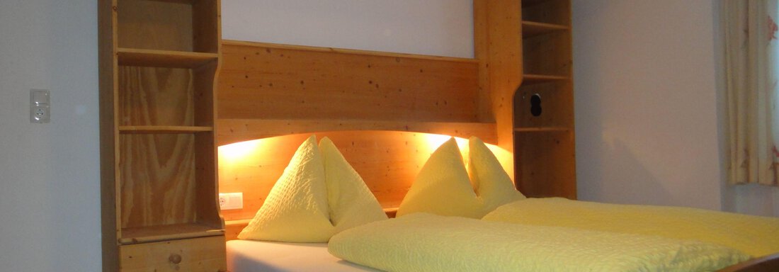 A cozy bedroom with a wooden bed and yellow pillows. There are shelves next to the bed and soft lighting. - Haus Elisabeth - Vent