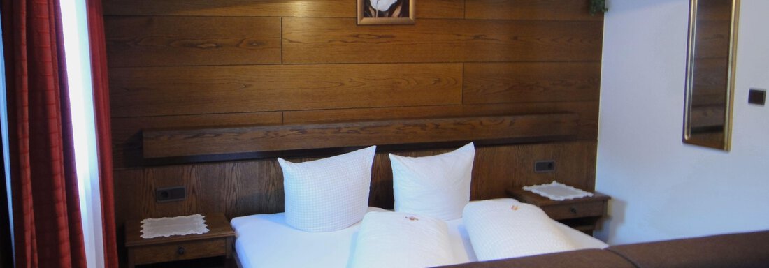 A cozy bedroom with wood paneling and two single beds. Next to the beds are bedside tables and a large window with red curtains. - Haus Elisabeth - Vent