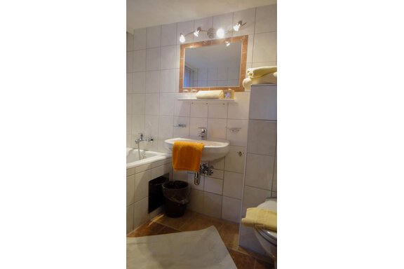 A modern bathroom with white tiles and a large mirror. There is a bathtub, a sink, and towels in shades of yellow. - Haus Elisabeth - Vent