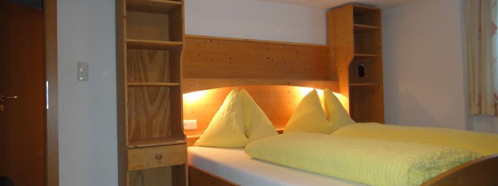 A cozy bedroom with a double bed and yellow cushions. The wooden shelves on the wall and the soft light create an inviting atmosphere. - Haus Elisabeth - Vent