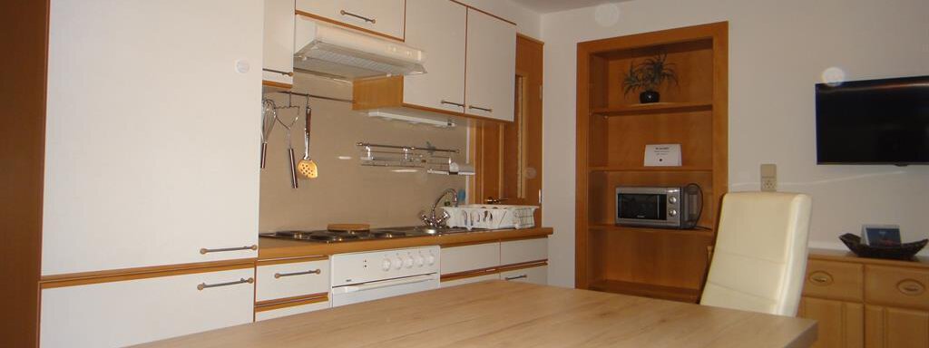 A modern kitchen with white cabinets and wooden shelves. The large table invites for a cozy meal. - Haus Elisabeth - Vent